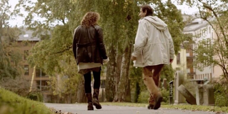 Due persone camminano lungo un sentiero di un parco alberato, con edifici visibili sullo sfondo.