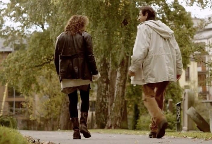 Due persone camminano lungo un sentiero di un parco alberato, con edifici visibili sullo sfondo. {{brizy_dc_image_alt entityId=